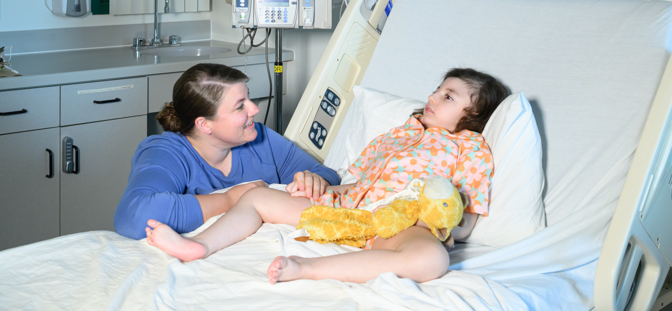 A mom sits beside her young daughter, who is in a hospital bed with a stuffed giraffe in her lap. They holding hands and facing each other.