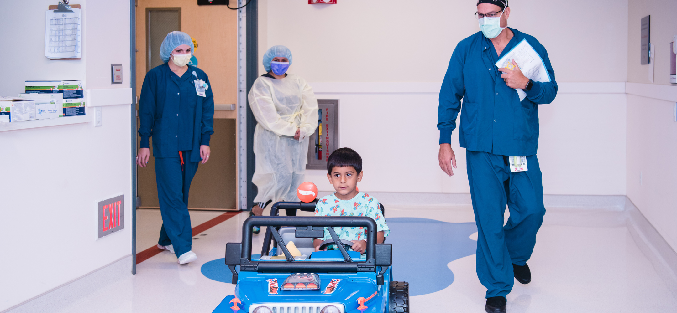 A patient drives a child-sized vehicle towards the camera, followed by an anesthesiologist, his mom and a nurse, on his way to the operating room.