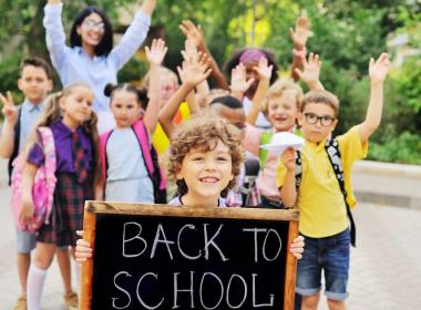 School age boy holds "Back to School" sign while friends and parents wave in the background